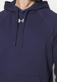 Personne portant un sweat à capuche bleu marine avec le logo Under Armour sur la poitrine et des cordons pendants.