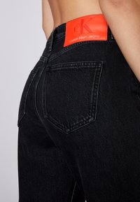 Svart denimjeans med en figurnära design, med två bakfickor och en framträdande orange Calvin Klein-logopatch på midjebandet.