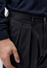 Svarta plisserade byxor med en mjuk tygtextur, med knäppning, dragkedja och en mörk midja, kombinerat med en marinblå tröja.