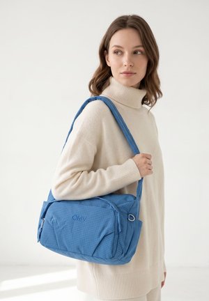 Donna con maglione beige a collo alto che porta una borsa a tracolla blu GMV, guarda di lato contro uno sfondo chiaro semplice.
