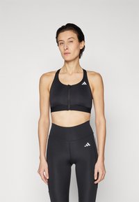 Soutien-gorge de sport noir avec fermeture éclair et bretelles larges, arborant le logo blanc d'Adidas. Associé à un legging noir taille haute avec accent logo.