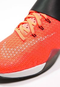 Scarpa da ginnastica di un vivace arancione con una tomaia testurizzata, dettagli neri e rifiniture bianche riflettenti. Presenta lacci arancioni e una suola in gomma.