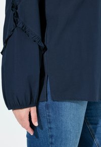 Marineblaues Oberteil mit Rüschendetail an den Ärmeln, mit lockerer Passform und seitlichen Schlitzen. Kombiniert mit hellblauen Jeans. Weiche Textur.