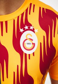 Maglia da calcio del Galatasaray con una base gialla e motivi rossi a forma di fiamma, un logo bianco con "1905" e cinque stelle sopra.
