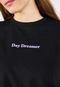 Sort bomulds T-shirt med et lilla "Day Dreamer" grafisk tryk. Klassisk rund halsudskæring, afslappet pasform, glat tekstur.