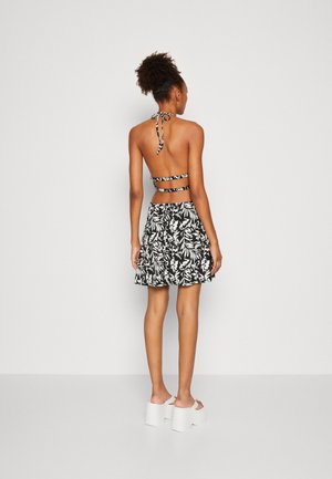 Femme aux cheveux bouclés portant une robe à fleurs noire et blanche avec un col halter et des sandales compensées blanches, debout devant un fond clair uni.
