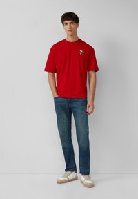 Rotes T-Shirt mit kurzen Ärmeln und einem kleinen Grafikdesign auf der Brust, kombiniert mit blauen Jeans und weißen Sneakers mit braunen Akzenten.