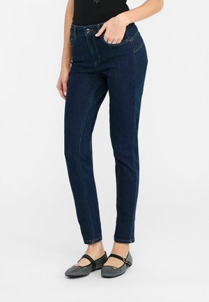 Dunkelblaue Skinny-Jeans aus Denim mit mittelhohem Bund, ausgestattet mit Fronttaschen und Kontrastnähten. Kombiniert mit schwarzen flachen Schuhen.