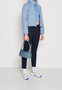 Osoba v světle modré péřové bundě, modrém zkráceném zipovém topu, tmavě modrých džínách, bílých teniskách a držící malé modré kabelky.