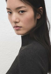 Close-up of a face with straight dark hair, neutral expression, wearing a dark ribbed turtleneck sweater against a light textured background.