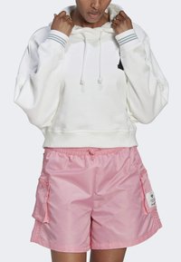 Sudadera blanca corta con rayas grises en los puños, combinada con pantalones cortos de cintura elástica rosa claro que cuentan con bolsillos y un parche con logo.