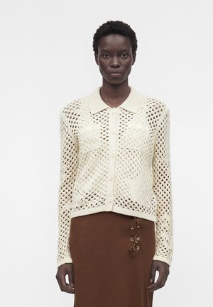 Woman wearing a cream crochet button-up cardigan with collar over a brown skirt featuring side lace-up details, standing against a plain background.