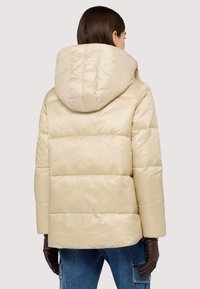 Béžová péřová bunda s velkou polstrovanou kapucí, prošívanou texturou a mírně zkráceným střihem. V kombinaci s tmavě hnědými rukavicemi.