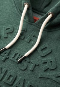 Sudadera de color verde oscuro con letras texturizadas, cordones blancos con extremos metálicos y un material similar a forro polar.