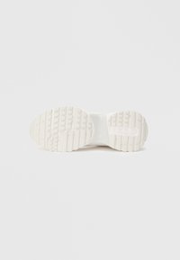 Hvit sneakersåle med teksturert gummi-mønster, med riller og mønstre for grep. Glatt mellomsåle som forbinder med overdelen.