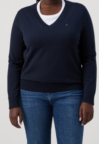 Maglione a V blu navy in materiale morbido, con maniche lunghe e polsini e orlo a coste. Abbinato a una maglietta bianca sotto, indossato con jeans blu.