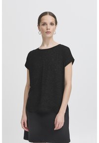 Naine, kellel on tagasisidetud juuksed, kuldsed rõngakõrvarõngad, lühikeste varrukatega must top ja must seelik, heleda ühevärvilise tausta ees.