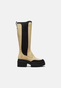 Botte haute en cuir beige avec panneau extensible noir, semelle épaisse à crampons noire et bout rond, vue de profil sur fond blanc.