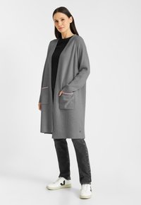 Grijze gebreide cardigan met open voorkant, geribbelde textuur, twee voorzakken met roze accent, gecombineerd met een zwarte top en donkere jeans.