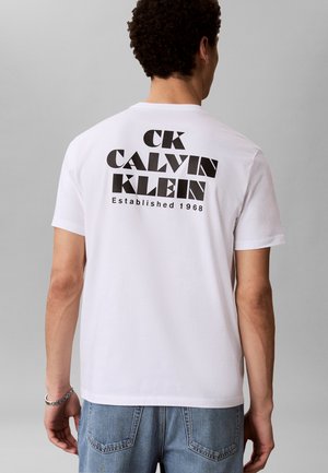 Muž nosící bílé tričko s krátkým rukávem s nápisem "CK CALVIN KLEIN Established 1968" velkými černými písmeny na zádech.