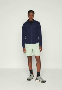 Giacca in pile con zip di colore navy sopra una maglietta nera, abbinata a pantaloni cargo verdi chiari. Indossata con calze nere e scarpe da corsa grigie.