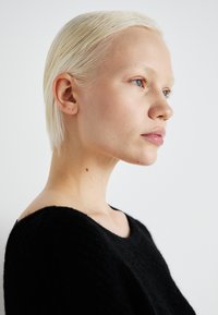 Close-up of a person with short, straight, platinum blonde hair wearing a black, textured sweater. Subtle freckles on the skin are visible.