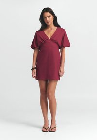 Bordeaux lyhyt mekko, jossa on syvä V-pääntie, puffihihat ja kerätty yksityiskohta edessä. Sileä kangas; malli käyttää yhteensopivia flip-flop-sandaaleja.