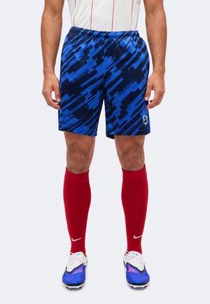 Homme portant un short de football bleu et noir à motifs, des chaussettes rouges montantes avec des logos blancs Nike, et des crampons bleus et blancs.