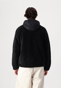 Giacca in pile nera con una superficie texturizzata e soffice, dotata di un cappuccio in nylon nero a contrasto. Vestibilità ampia con maniche lunghe e senza chiusure visibili.