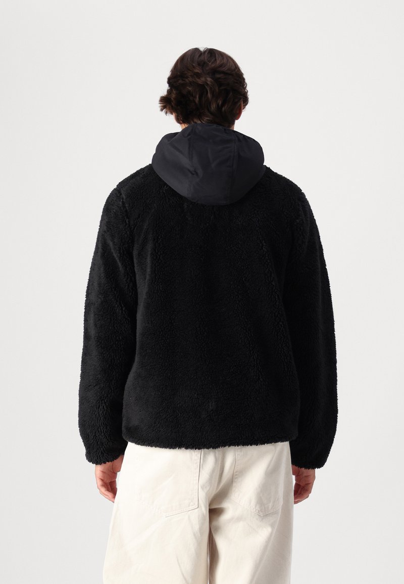 Chaqueta de forro polar negra con una superficie texturizada y esponjosa, y una capucha de nailon negro contrastante. Ajuste holgado con mangas largas y sin cierres visibles.