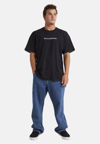 Zwarte katoenen t-shirt met korte mouwen en wit logo. Gecombineerd met losse blauwe denim jeans en zwarte instappers. Eenvoudig en casual ontwerp.