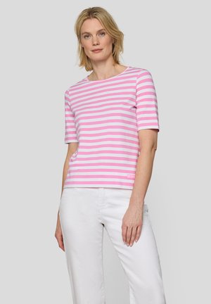 Frau trägt ein rosa-weiß gestreiftes T-Shirt mit kurzen Ärmeln und weiße Hose, steht vor einem einfarbigen hellen Hintergrund.