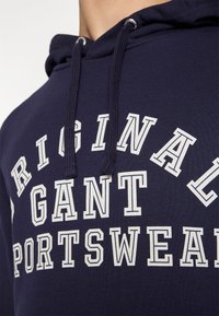 Sweat à capuche bleu marine avec texte blanc imprimé indiquant "ORIGINAL GANT SPORTSWEAR", doté d'un cordon de serrage et d'une texture côtelée au niveau du col.