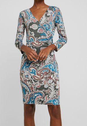 Nainen pukeutuneena pitkähihaiseen, polvipituiseen kietaisumekkoon, jossa on sinisiä, valkoisia ja ruskeita paisley-kuvioita tummalla taustalla.