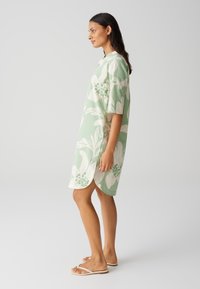 Short-sleeve, knee-length dress in light green fabric with large white floral patterns and green accents, featuring side slits and a relaxed fit.