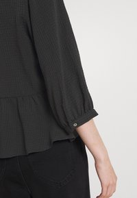 Blusa grigia scura con texture, maniche lunghe e svasate, orlo arricciato e bottoni decorativi sui polsini. Abbinata a pantaloni neri.