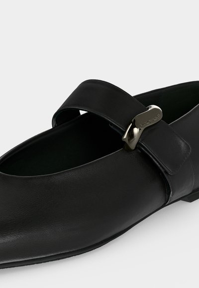 Mocassin en cuir noir à enfiler avec une large bride et un détail de boucle métallique argentée sur un fond clair.