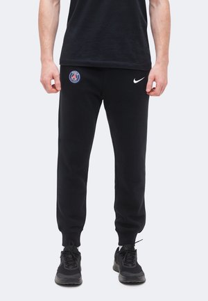 Homme portant un pantalon de survêtement noir avec les logos du Paris Saint-Germain et de Nike, assorti à des baskets noires et un t-shirt noir.