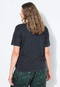 Kvinde med bølget brunt hår iført en falmet sort t-shirt og bukser med grøn paisley-mønster, der står til venstre foran en ensfarvet hvid baggrund.