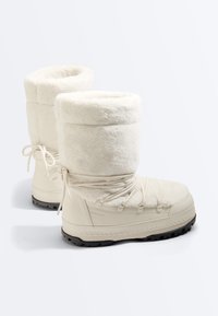 Paire de bottes d'hiver couleur crème mi-mollet avec une doublure en fausse fourrure, des lacets en tour de cheville et des semelles noires texturées pour l'adhérence.