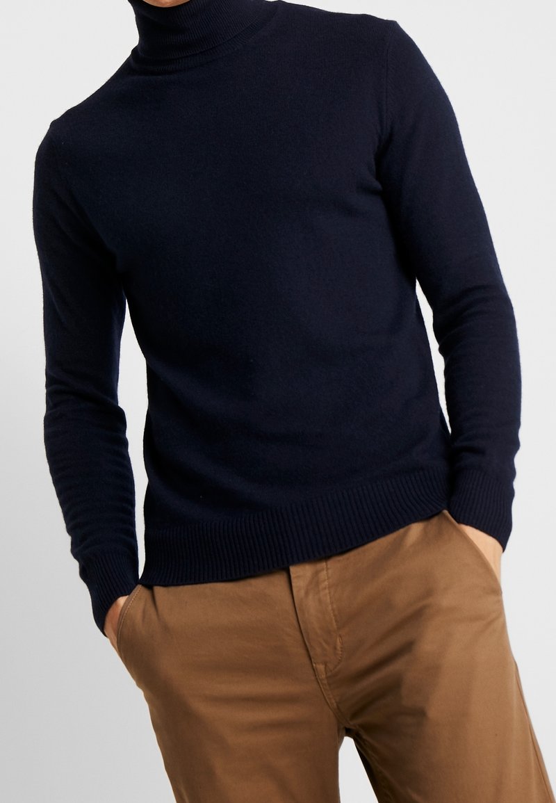 Marineblauwe gebreide turtleneck trui met geribde cuffs en zoom, gemaakt van zacht materiaal. Gecombineerd met lichtbruine chinos.