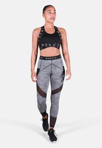 Czarny stanik sportowy z logo, w połączeniu z szarymi legginsami z siateczkowymi panelami i geometrycznymi wzorami. Model nosi czarne buty sportowe.