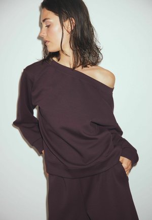 Mørkelilla oversized sweatshirts med asymmetrisk halsudskæring, lange ærmer og en afslappet pasform. Stoffet ser blødt og glat ud.