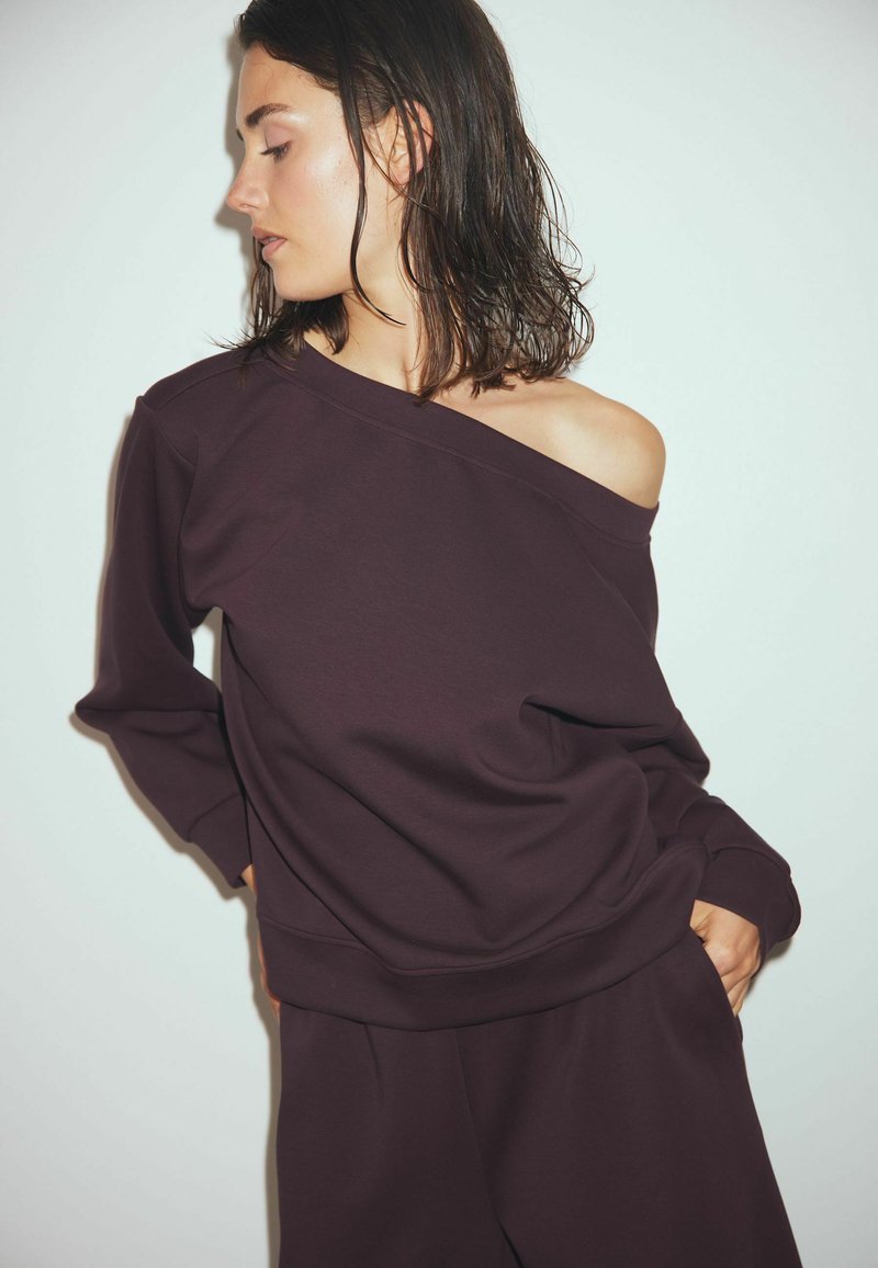 Dark purple oversized sweatshirt with an asymmetrical neckline, long sleeves, and a relaxed fit. The fabric appears soft and smooth.