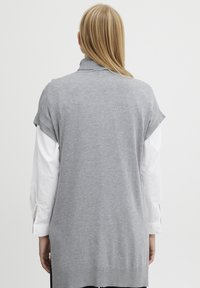 Grijze mouwloze trui met een rolkraag, gemaakt van zachte stof, met een losse pasvorm en zijsplitten, gedragen over een witte lange mouw shirt.