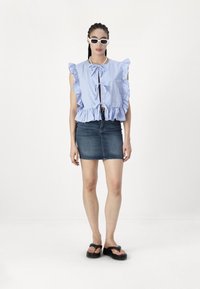 Blouse à rayures bleues et blanches avec volants et nouage à l'avant, associée à une mini jupe en denim foncé et des tongs noires. Lunettes de soleil blanches portées.