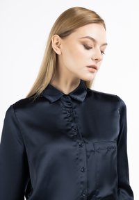 Camicia di satin blu navy con colletto, bottoni frontali e tasca sul petto. Il tessuto appare liscio con una finitura lucida e minima texture.