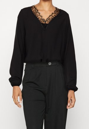 Zwarte blouse met lange mouwen en kanten V-halsdetail, ingestopt in hooggetailleerde zwarte broek met voorste knoop en riemlussen.