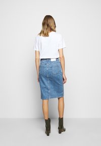 Woman standing facing away, wearing a white t-shirt, knee-length blue denim skirt, and olive green ankle boots with block heels.