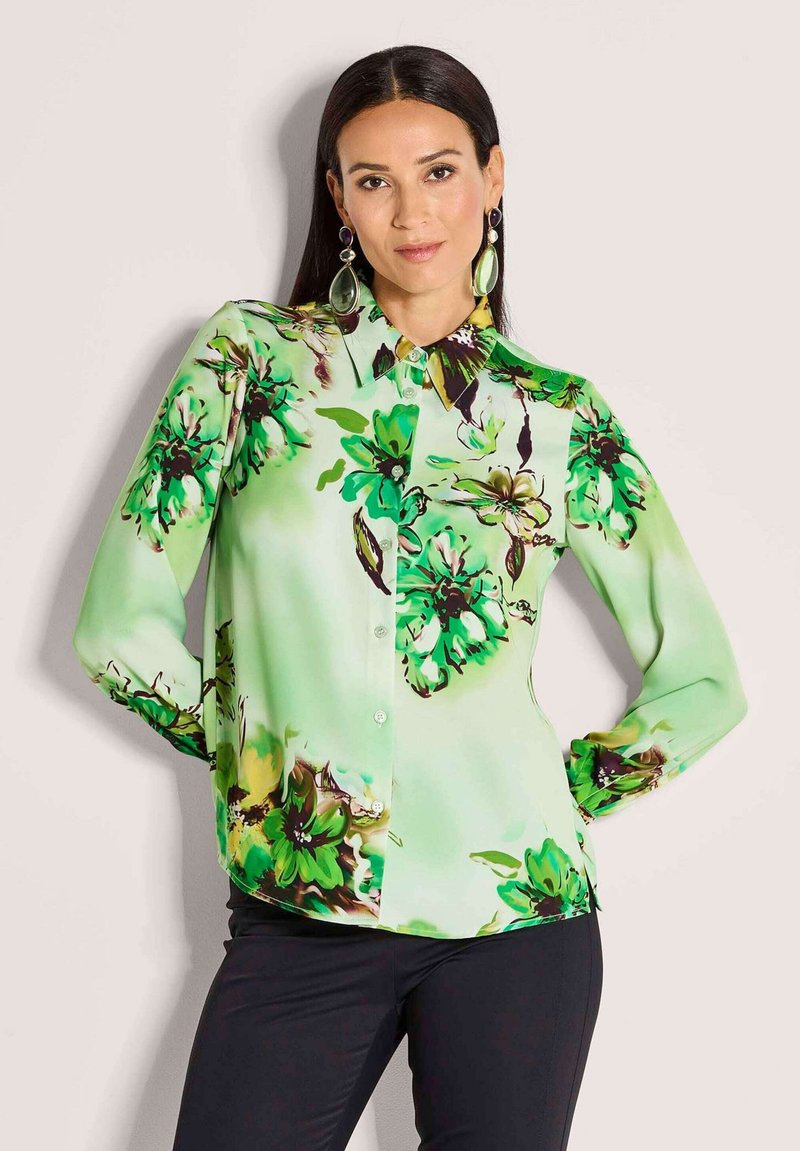 Vrouw met lang donker haar draagt een groene bloemetjesblouse met knopen en grote groene hangende oorbellen, poseert met haar handen op haar heupen.